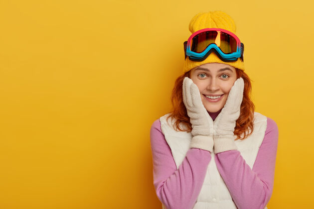 红发可爱快乐的女人双手放在脸颊上 温柔的微笑 微笑着看着镜头 享受着积极的休息和滑雪旅游 穿着积极的衣服 隔离在黄色的墙壁上冬天的时间冬天触摸交叉