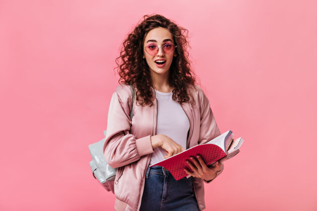 生活方式一个穿着粉色夹克和牛仔裤的惊讶女人在孤立的背景下和书合影笔记本毛衣黑发