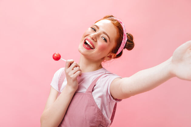 头带穿着粉色连身衣和白色上衣的快乐女人拿着糖果 在粉色背景上自拍女人化妆红发