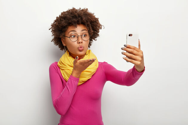 设备可爱的非洲裔美国女人的肖像送空气吻 通过智能手机自拍 戴透明眼镜 做圆嘴唇 穿生动的衣服 隔离在白色的墙壁上自拍Poloneck眼镜