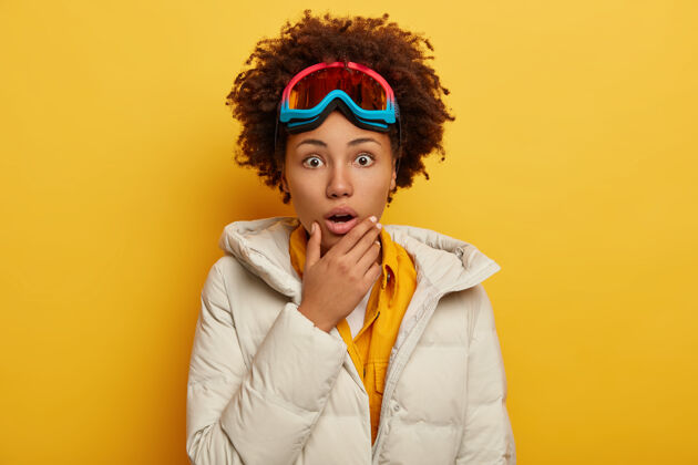 寒冷人 情感 爱好和休闲的概念惊恐情绪卷曲的非洲裔美国妇女戴着滑雪板面具 穿着蓬松的白大褂 托着下巴夹克冬天黄色