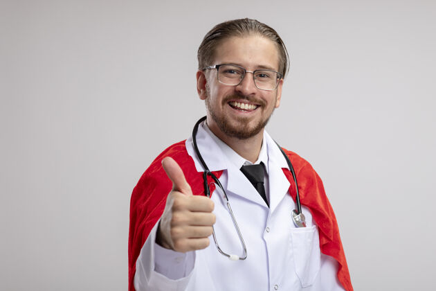 眼镜年轻的超级英雄微笑着穿着医用长袍 戴着听诊器和眼镜 大拇指朝上年轻家伙微笑