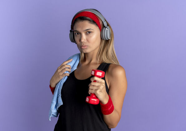 耳机身穿黑色运动服 头戴红色耳机 手持哑铃 表情严肃的年轻健身女孩站在蓝色的墙上运动装脸年轻