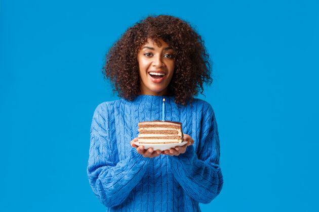 乐趣开朗而梦幻的可爱的非洲裔美国b-day女孩 拿着蜡烛拿着蛋糕 吹着蜡烛笑着 开着生日派对 站在毛衣蓝色的墙上女性年轻肖像