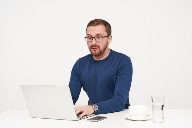 坐着困惑的年轻金发男子戴着眼镜 手放在键盘上 坐在白色背景上惊奇地看着笔记本电脑的屏幕水套头衫蓝色