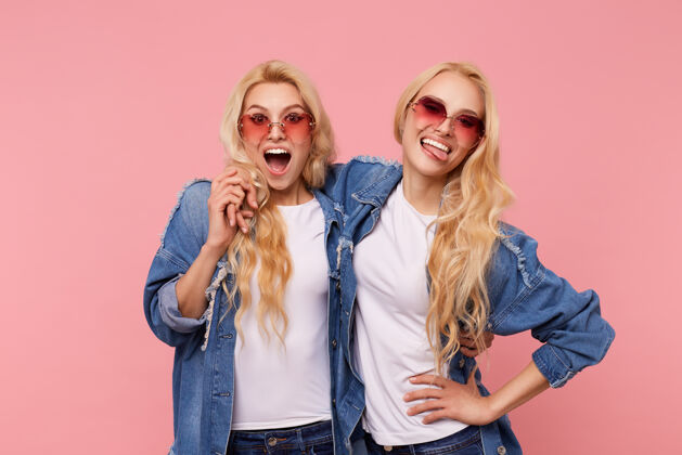 粉色年轻可爱的金发双胞胎的室内照片 波浪形的发型相互拥抱 一边笑一边玩 穿着休闲服站在粉色背景上室内积极卷发