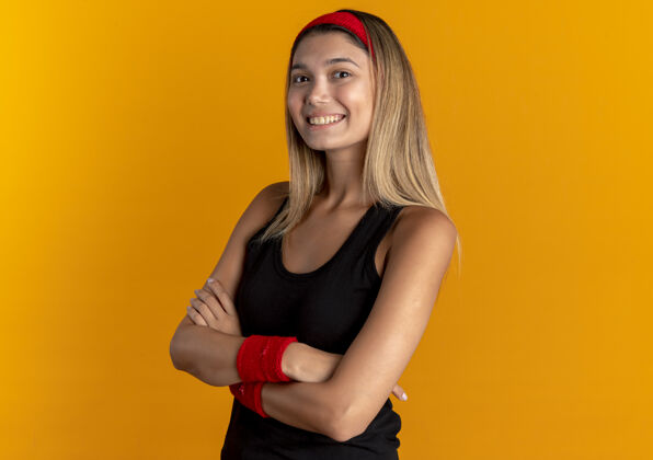 运动装身穿黑色运动服 头戴红色头巾的年轻健身女孩 双臂交叉 站在橙色的墙上 看上去自信满面自信健康十字架