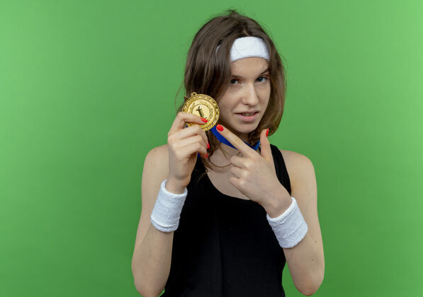 手指身穿黑色运动服的年轻健身女孩戴着头带 脖子上挂着金牌 手指指着它 站在绿色的墙上 显得很自信站奖牌运动装
