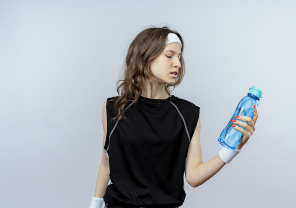 抱着一个穿着黑色运动服的年轻健身女孩 戴着头巾 手里拿着一瓶水 严肃地站在白色的墙上看着它看着女孩站着