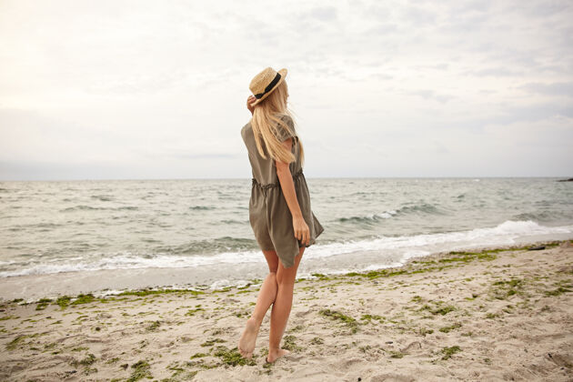 绿色一张年轻苗条的长发金发女郎的户外照片 她穿着绿色的夏装 背对着海滩 手放在帽子上波浪后面风