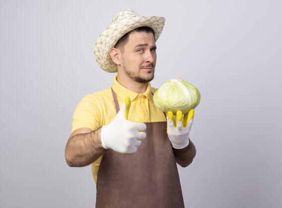 卷心菜年轻的园丁穿着连体衣戴着帽子戴着工作手套拿着卷心菜看着摄像机竖起大拇指拇指男人向上
