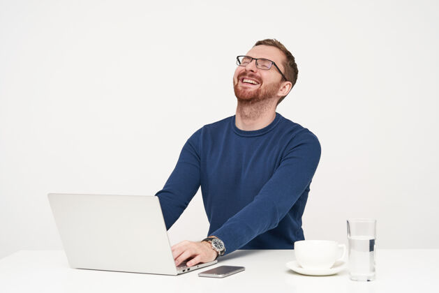 毛衣快乐的年轻胡须金发男子抛回他的头 同时笑 眼睛闭上 听到有趣的笑话 而他的笔记本电脑在白色背景下工作坐着男性眼镜