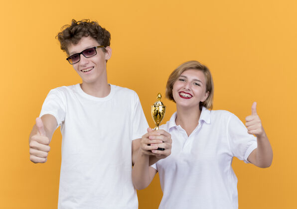 情侣年轻的运动情侣站在一起举着奖杯 微笑着竖起大拇指站在橙色的墙上女人表演抱着