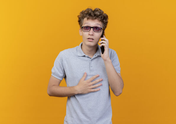 手机一个戴着黑眼镜 穿着灰色马球衫的年轻人站在橘色的墙上讲着手机 看上去很惊讶惊喜说话穿