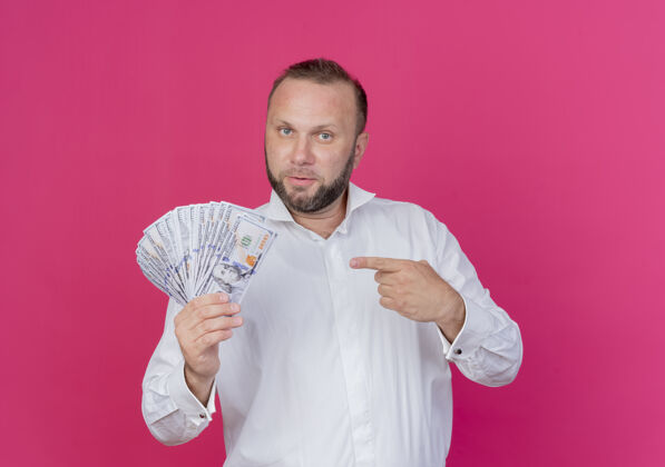 男人一个留着胡子的男人穿着白衬衫 用食指指着钞票 站在粉色的墙上 看起来很自信现金胡须指向