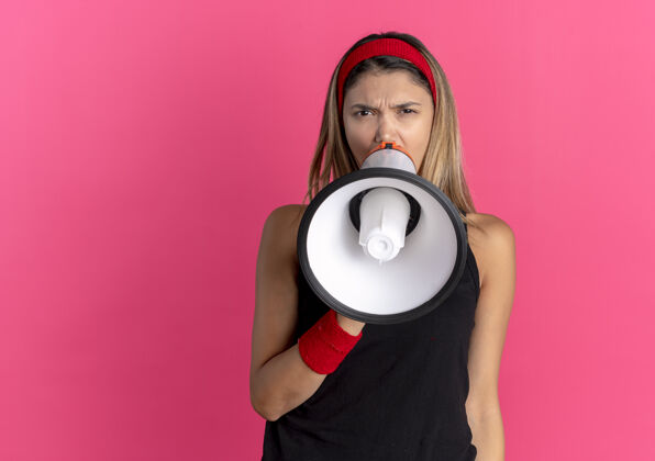 运动装身穿黑色运动服 头戴红色头巾的年轻健身女孩对着站在粉色墙上的扩音器大声喊叫女年轻私人教练