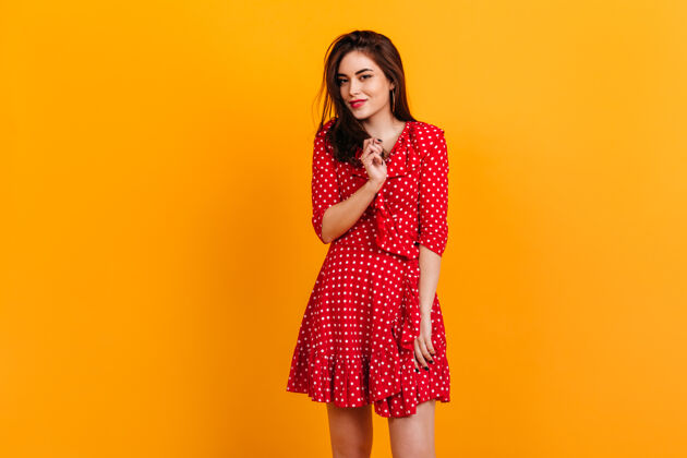 站立穿着红色裙子的时髦女孩的肖像模特在橙色的墙上微笑着黑发颜色头发