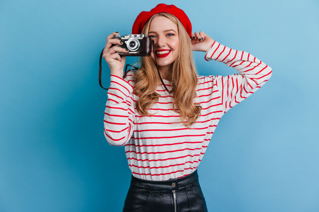 帽子穿着条纹衬衫的快乐女孩拿着相机法国女模特在蓝色墙上拍照欢呼卷发法国