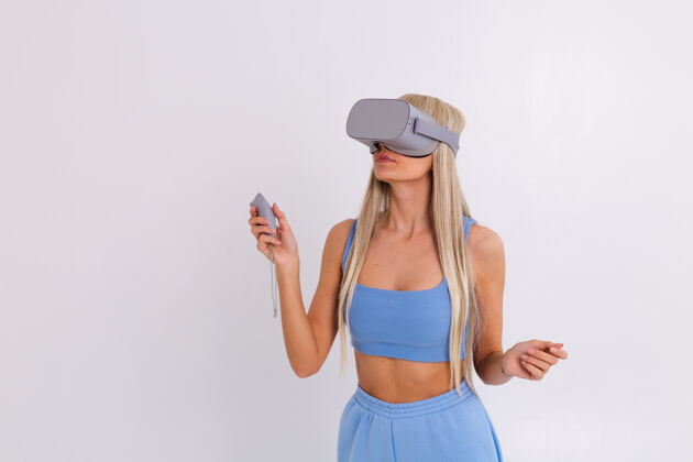 姿势摄影棚照片中一个年轻迷人的女子穿着温暖的蓝色时尚西装 戴着虚拟现实眼镜在白色的设备表情游戏