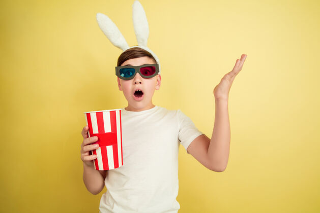 肖像戴着爆米花的眼镜看电影白人男孩在黄色背景上扮演复活节兔子复活节快乐漫画面部惊喜