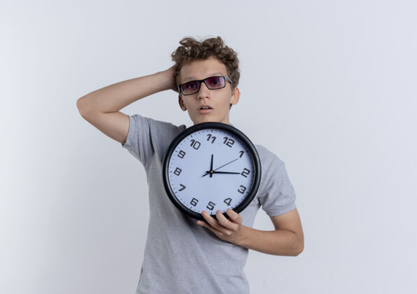 戴着戴着黑眼镜的年轻人穿着灰色马球衫 手里拿着挂钟 头上的手站在白色的墙上眼镜人马球