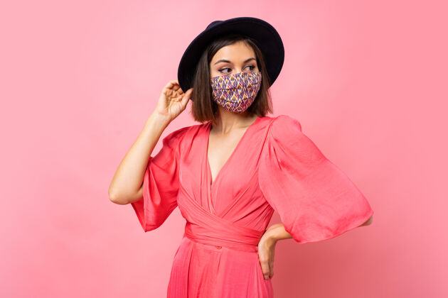 阳性时髦的女人戴着保护性的时髦的面具戴着黑色的帽子和太阳镜在粉红色的墙上摆姿势社交学生特写