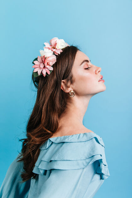 欢呼身着褶边衬衫的贵族女孩的侧面照片头发上插着鲜花的女士骄傲地在蓝色的墙上摆姿势成人魅力粉色花朵