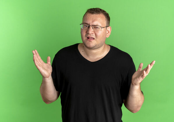 耸肩一个戴眼镜的胖男人穿着黑色t恤 看起来很困惑 耸耸肩站在绿色的墙上眼镜超重男人