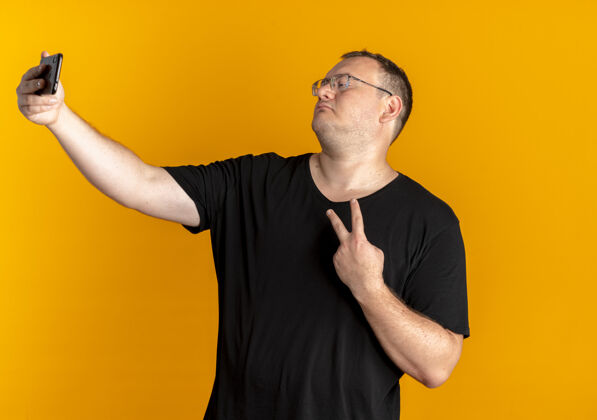 眼镜戴眼镜的超重男子身穿黑色t恤手持智能手机自拍显示胜利标志站在橙色的墙上展示站着抱着