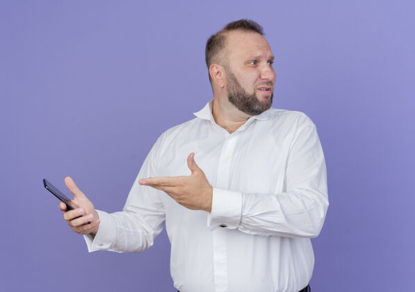 抱着一个留着胡子的男人穿着白衬衫 手拿着智能手机 站在蓝色的墙上 手臂朝一边看移动胡须站着