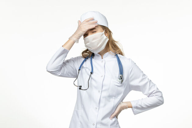 女性正面图年轻女医生穿着医疗服戴着防护口罩由于冠状病毒在浅白色的表面冠状病毒疾病套装