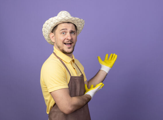 胳膊年轻的园丁 戴着工作手套 穿着连体衣 戴着帽子 手上拿着东西 高兴地笑着看紫色连身衣