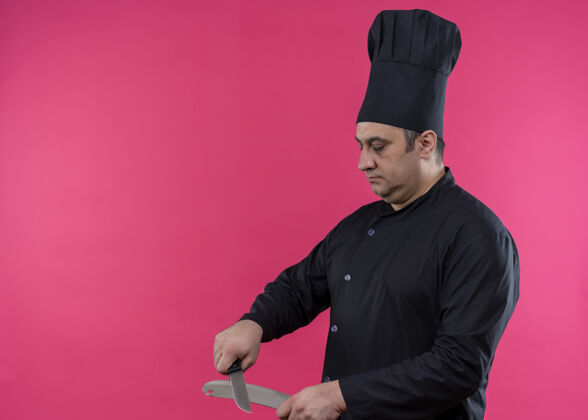 磨刀器男厨师身穿黑色制服 头戴厨师帽 手持削尖刀 表情严肃 站在粉色背景上烹饪刀粉色