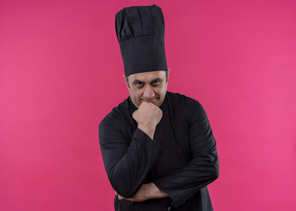 看体贴的男厨师身穿黑色制服 头戴厨师帽 手放在下巴上 站在粉色背景上沉思地看着镜头帽子粉色穿