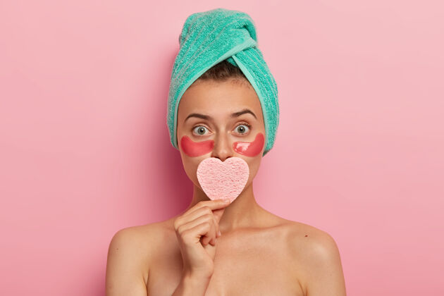 海绵迷人的女人有最少的化妆品 在眼睛下面涂化妆海绵 用心形海绵盖住嘴 有完美的护理皮肤 看起来出奇 赤裸的身体站在室内治疗删除自然