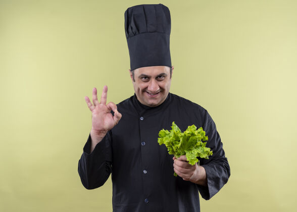 帽子男厨师身穿黑色制服 头戴厨师帽 手拿新鲜生菜 微笑着 绿色背景上立着ok标志站着标志穿上