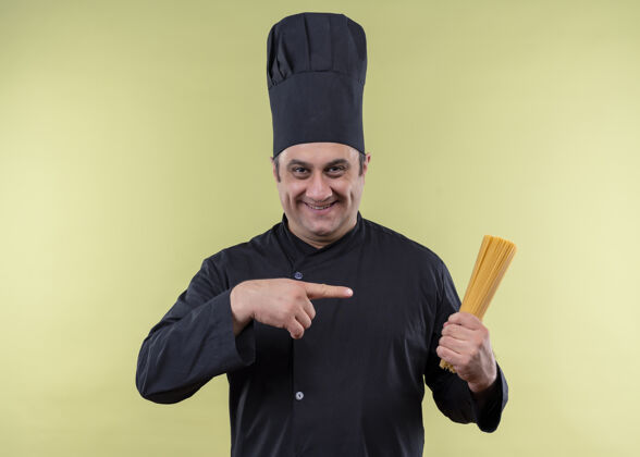 厨师男厨师身穿黑色制服 头戴厨师帽 手拿一排意大利面 手指指着它 看着摄像机 站在绿色背景下 满脸笑容站着抱着手指