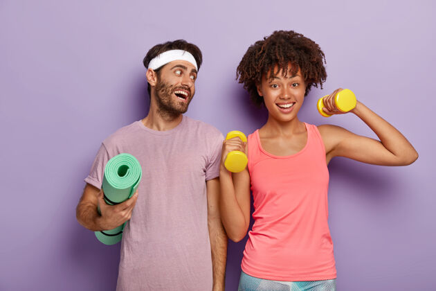 微笑积极的运动员戴着头带和t恤 拿着皱巴巴的健身垫 高兴地看着举着哑铃的女友 一起锻炼女朋友一起快乐