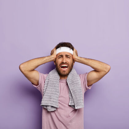 健身房不满意的未刮胡子的男人手放在头上 头痛 在室内锻炼 穿着休闲t恤 肩上搭毛巾 训练后筋疲力尽 精神低落胡茬穿姿势