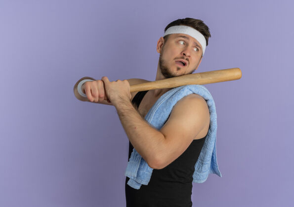 自信戴着头带 肩上扛着毛巾 手持棒球棒 自信地站在紫色背景下 一边看一边的年轻健身男子球棒肩膀健身