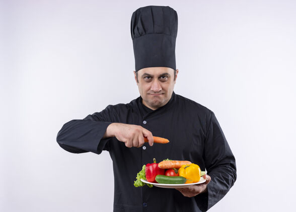 严肃男厨师身穿黑色制服 头戴厨师帽 拿着盛有新鲜蔬菜和胡萝卜的盘子 站在白色背景上严肃地看着摄像机拿着看蔬菜
