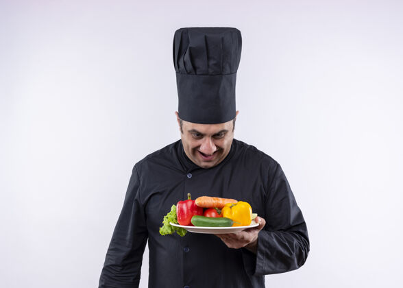 烹饪男厨师身穿黑色制服 头戴厨师帽 手持盛有新鲜蔬菜的盘子 面带微笑地看着盘子 站在白色背景上黑色厨师盘子