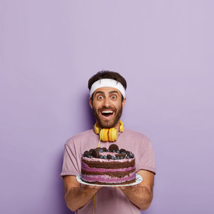 没刮胡子喜出望外的运动员手拿蓝莓烤制的大水果蛋糕 想在健身房里精疲力尽的运动后吃点甜食 穿着休闲服 戴着耳机听音乐训练男性健美操