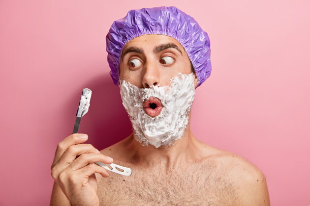 健康震惊的欧洲男人拿着剃须刀 在脸颊上涂上泡沫凝胶 剃毛 戴上紫色的保护性淋浴帽 有卫生习惯阳刚印象年轻