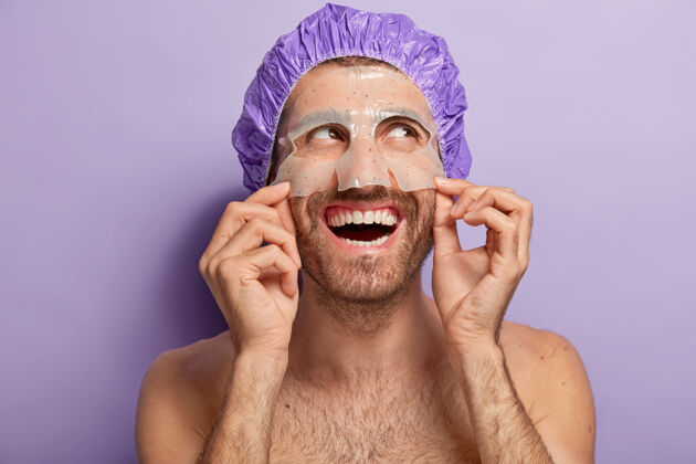 沙龙开朗的年轻人戴着保湿面膜 淋浴帽 赤身裸体站在室内 表情愉快 喜欢美容程序紫罗兰色干净水平
