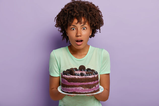 情绪照片中惊讶的黑皮肤女人卷发 捧着美味的蛋糕 惊讶的客人已经在门口吃饭 穿着休闲服 烤着美味的甜点胃口紫罗兰美味