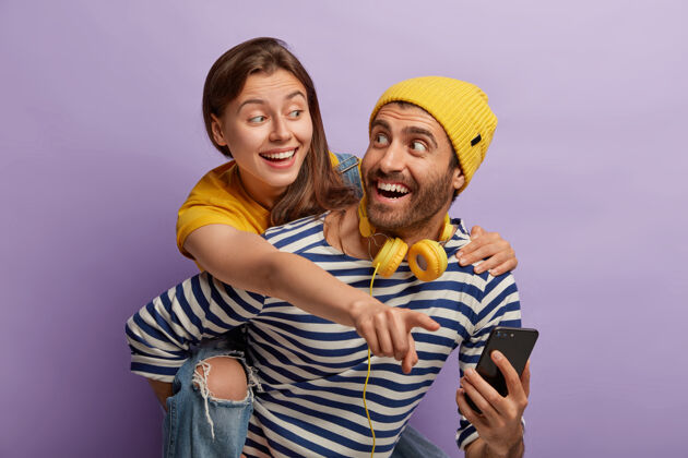 积极一对幸福的欧洲夫妇在一起玩得开心 用现代科技娱乐高兴的男人背着女朋友 戴着黄色帽子和条纹套头衫 拿着手机 展示图片乐趣家庭黄色