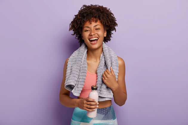 耐力无忧无虑的积极的非洲裔美国妇女穿着运动服 拿着一瓶淡水 高兴健康 有规律的训练 脖子上带着柔软的毛巾积极高兴健康