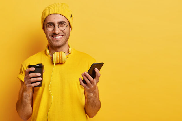 帽子帅气开朗的年轻人的画像 脸上洋溢着喜悦的表情 手持手机 给朋友发短信 喝外卖咖啡 戴着眼镜 黄色的行头戴着耳机肖像使用电话