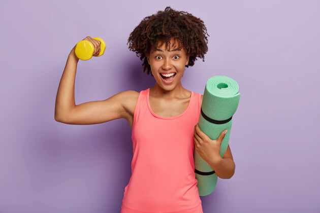 女子非洲发型快乐女人横拍 有肱二头肌举重 拎着卷起的健身垫 穿着粉色背心 神采奕奕 靠着紫色墙壁模特运动 动力形状欢呼私人教练